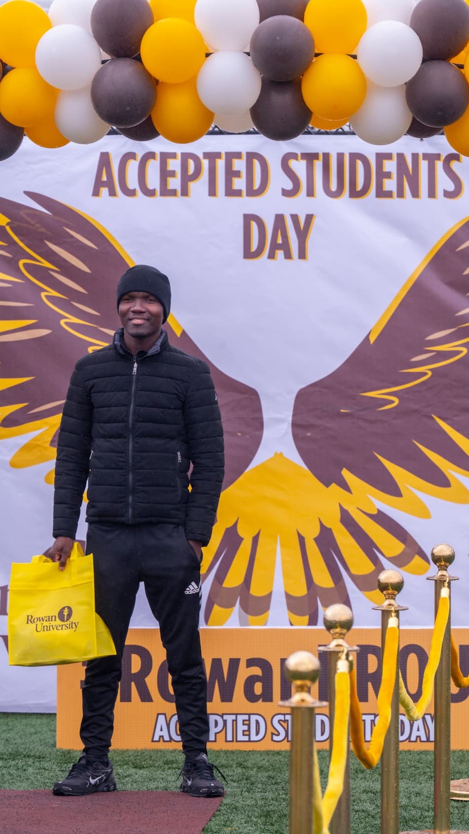 an accepted student wearing a black hat, jacket, and pants standing in front of an Accepted Students Day backdrop