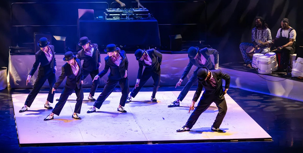 A light-colored tap floor center stage with 7 dancers all in the same pose with right leg and arm outstretched to the side, in black suits with black and white spectator shoes, behind them on a platform is a DJ at a turntable, stage left are 2 bucket drummers.