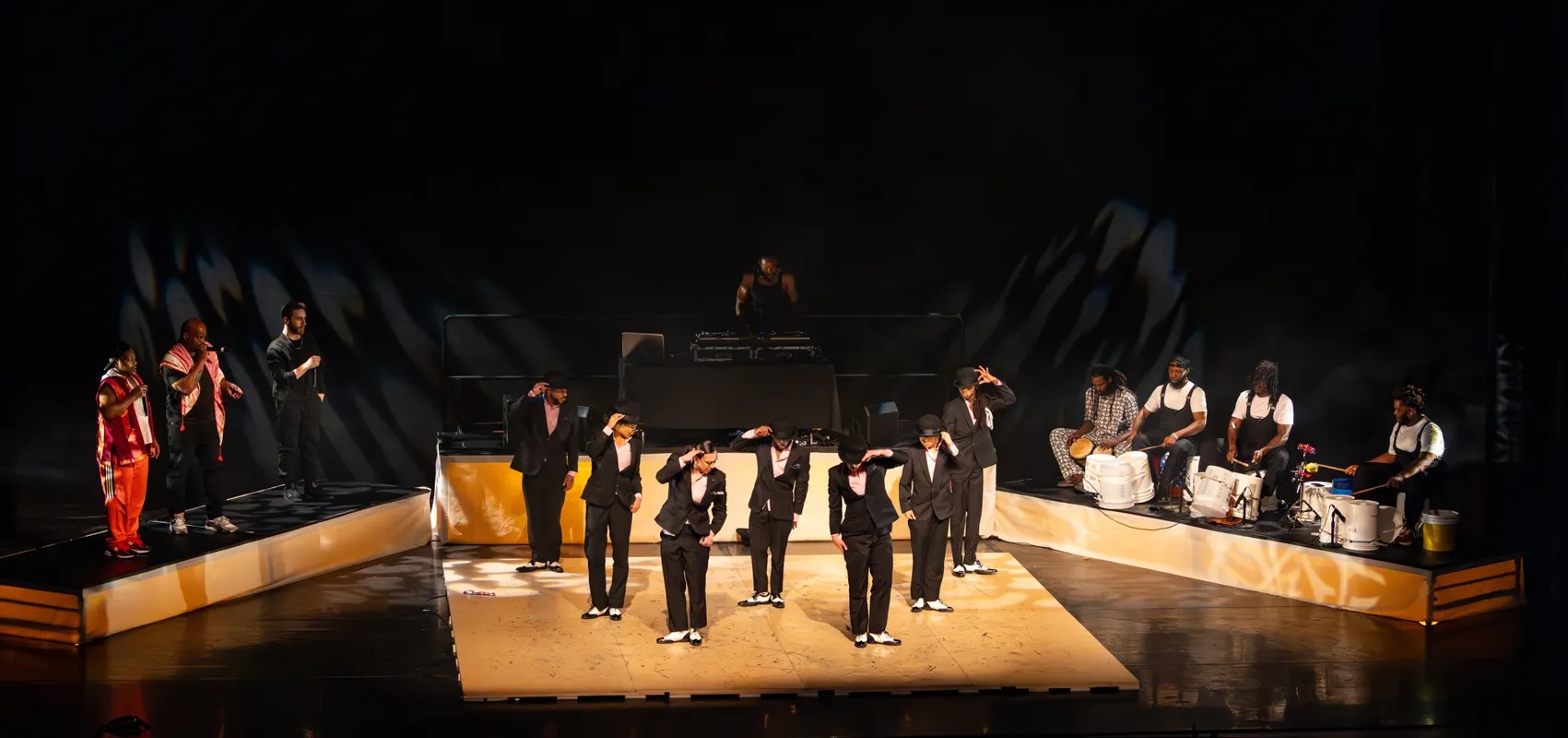 A light-colored tap floor center stage with 7 dancers in a V formation, in black suits with black and white spectator shoes, behind them on a platform is a DJ at a turntable, stage left are 4 bucket drummers, and stage right are 3 beat boxers holding microphones.