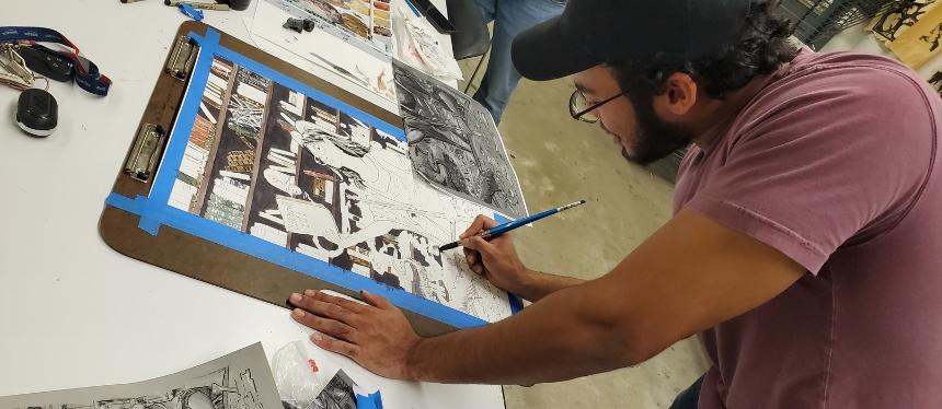 An artist in a red t-shirt and baseball cap works on an illustration over a lightbox.
