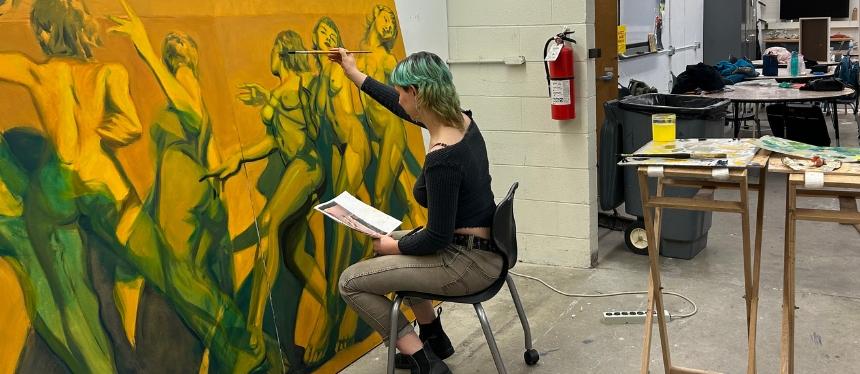 An artist sits before a large canvas working on a painting of nude women in motion. The artist holds a paintbrush up to the canvas with their right hand and a reference image in their left.