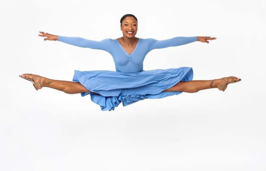 One Black female dancer mid-air facing the camera in a light blue dress against a white background, in a wide straddle, with both legs extended to either side and pointed toes.