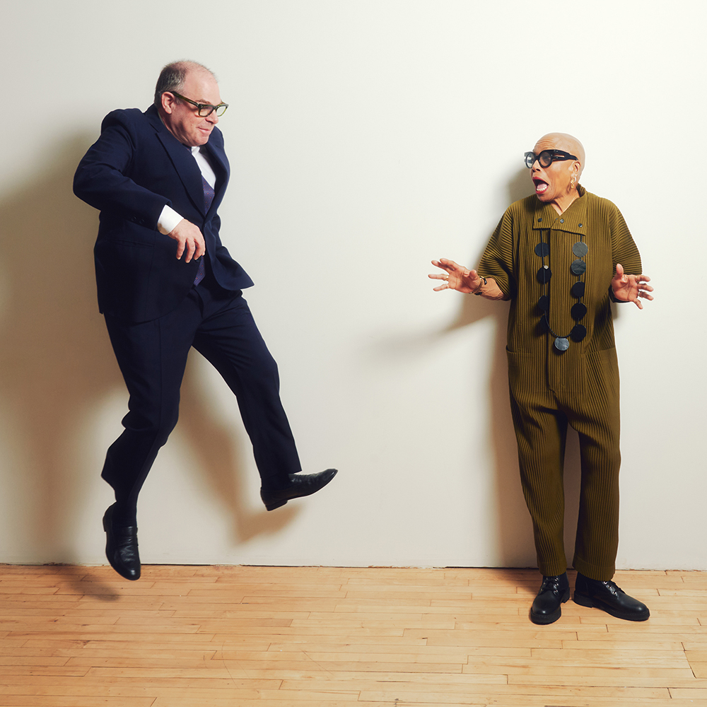A balding white man in blue suit jumping against a white wall, facing a bald Black woman looking scared, but jokingly, in an olive green shirt and pant set with big black glasses.