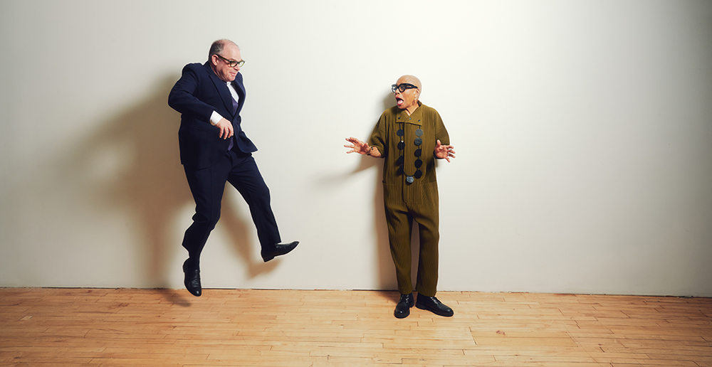 A balding white man in blue suit jumping against a white wall, facing a bald Black woman looking scared, but jokingly, in an olive green shirt and pant set with big black glasses.