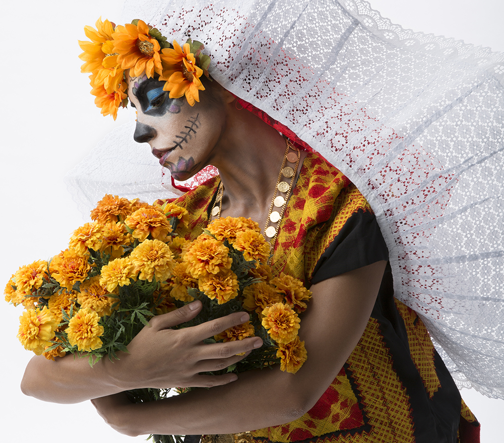 Closeup side profile of a woman with sugar skull face makeup, a marigold-lined white-lace veil, holding a bouquet of marigolds, which she is looking down upon, solemnly.