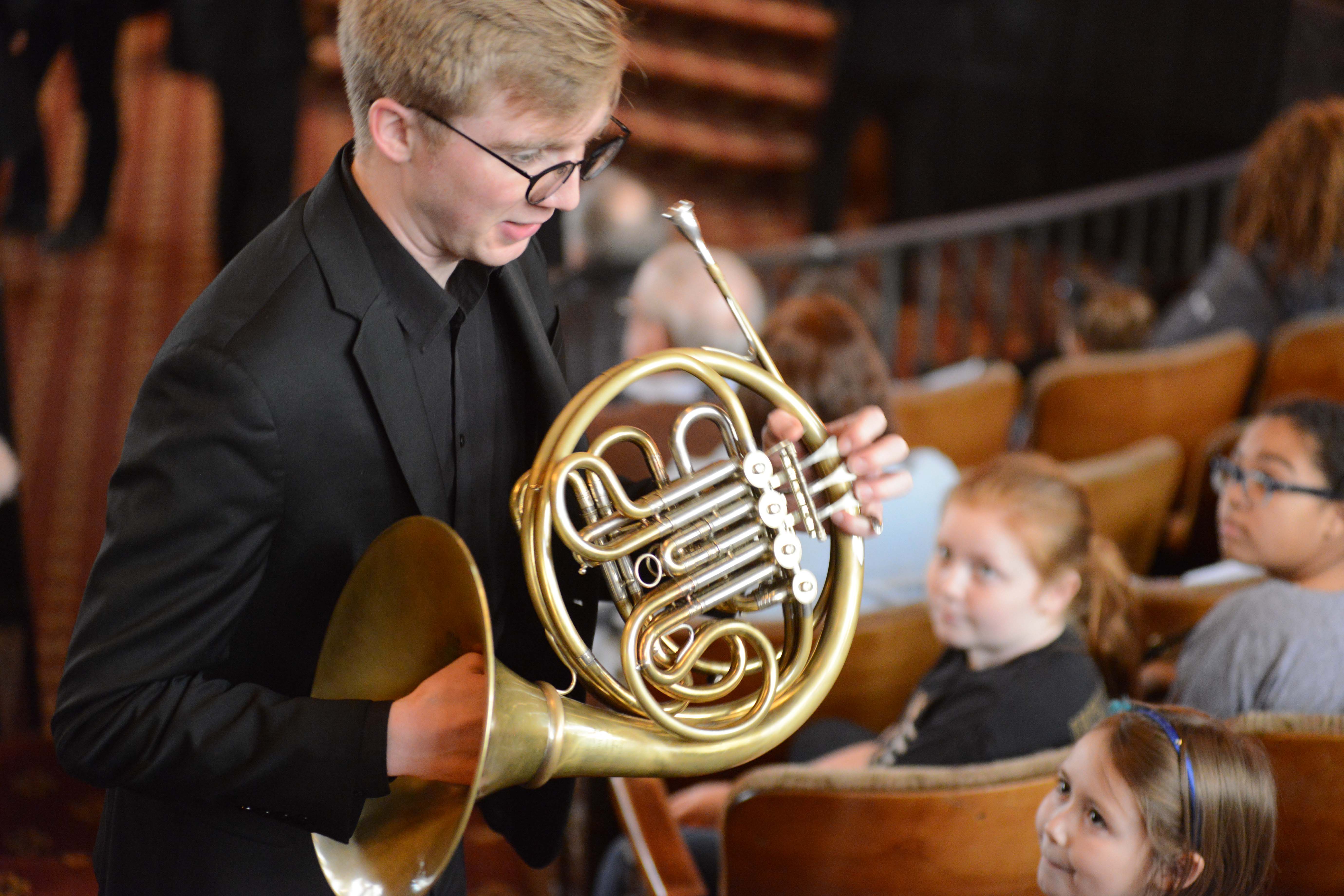 A musician holding a french horn walking through a crowd of children seated in an auditorium