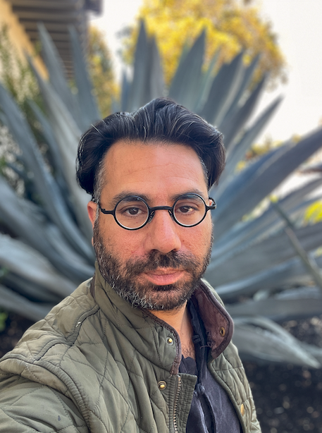 Samuel wearing a green quilted jacket and glasses, smiling at the camera