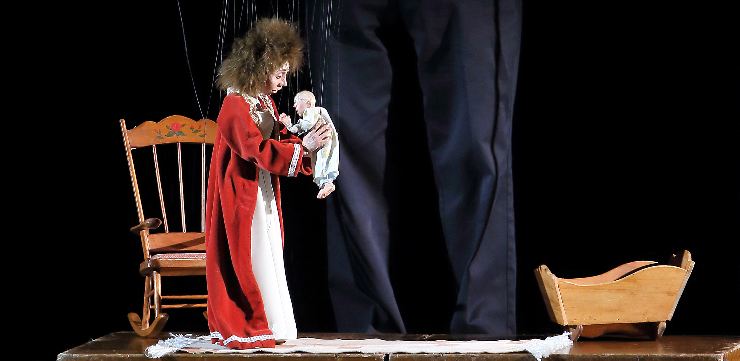 Marionette of a mother in a robe holding a baby near a basinnette rocker.