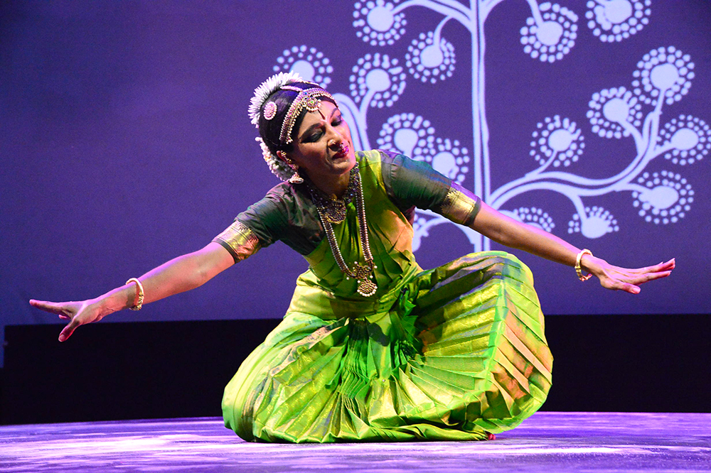 A seated Bharatanatyam dancer in bright green costume seated on the stage, bowing to the earth below.