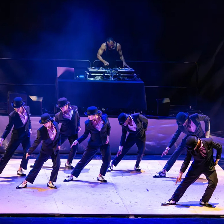 A light-colored tap floor center stage with 7 dancers in a V formation, in black suits with black and white spectator shoes, behind them on a platform is a DJ at a turntable.