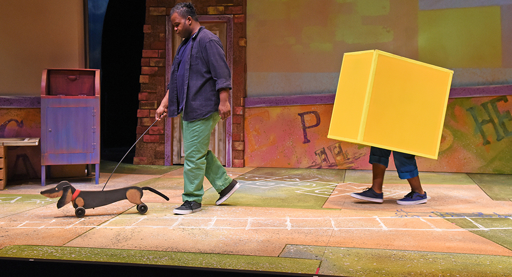 A Black man walking a wooden dachshund on wheels with a Black child hiding under a yellow box following behind him against a theatrical backdrop that looks like a city block.