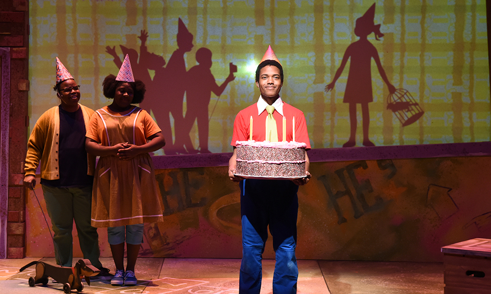 A Black boy center stage holding a birthday cake and wearing a paper birthday hat, with his parents grinning behind him and shadow puppets projected in the background as silhouettes of other party guests.