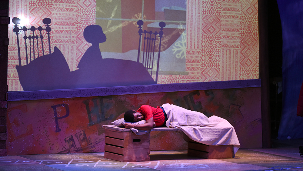 A Black boy sleeping on a bed on stage with a projected scene behind him of a silhouette of a boy sitting up in his bed looking out his window to a snowy scene.