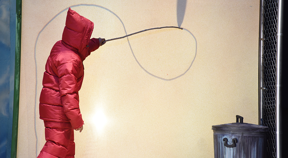 A boy in a red snow suit drawing a figure eight pattern in the snow with a stick.