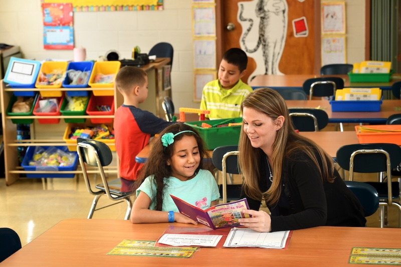 A teacher works one-on-one with a students while other students sit in the background.