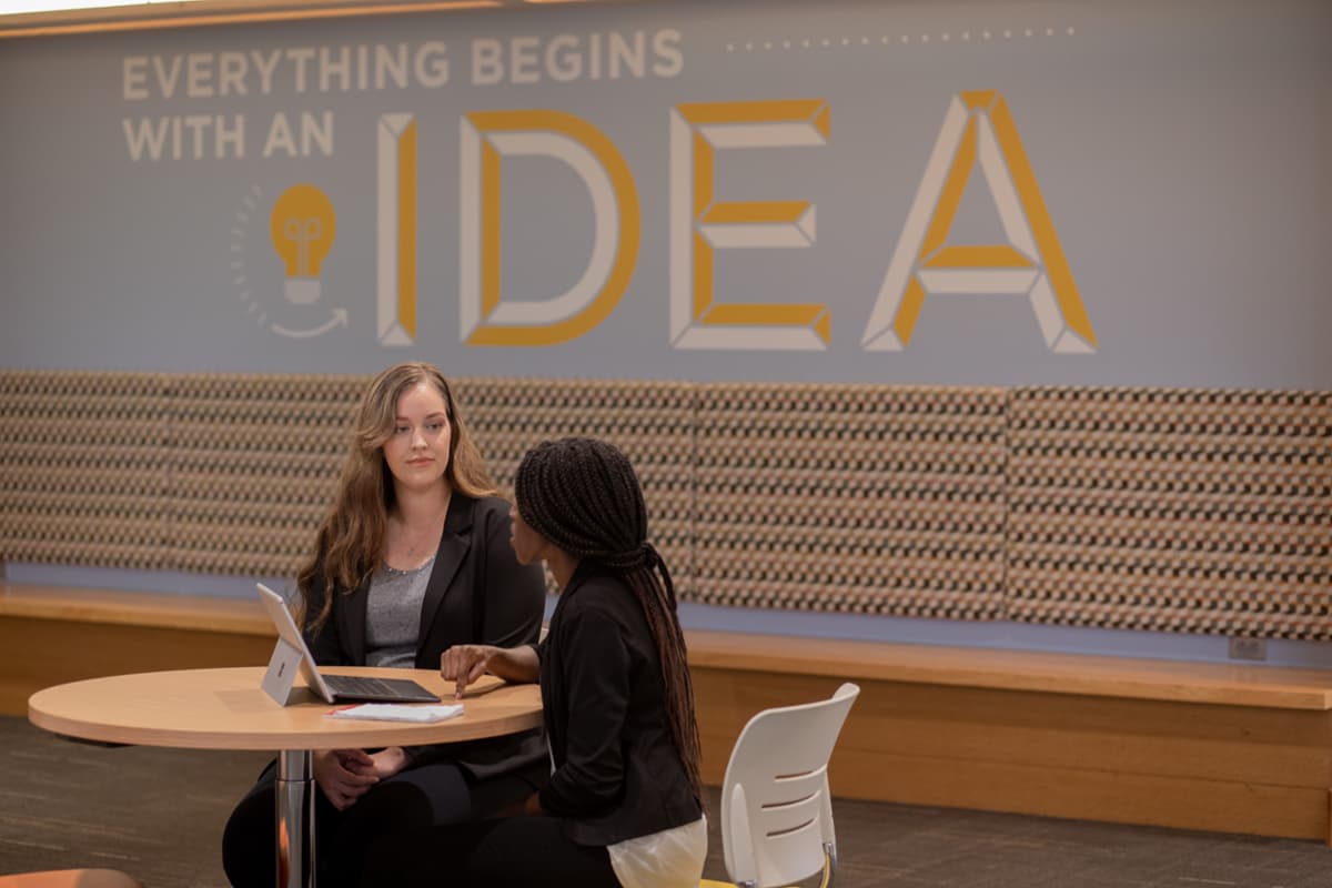 A Rowan student talking with a staff member in front of a wall that says Everything Begins with an Idea.