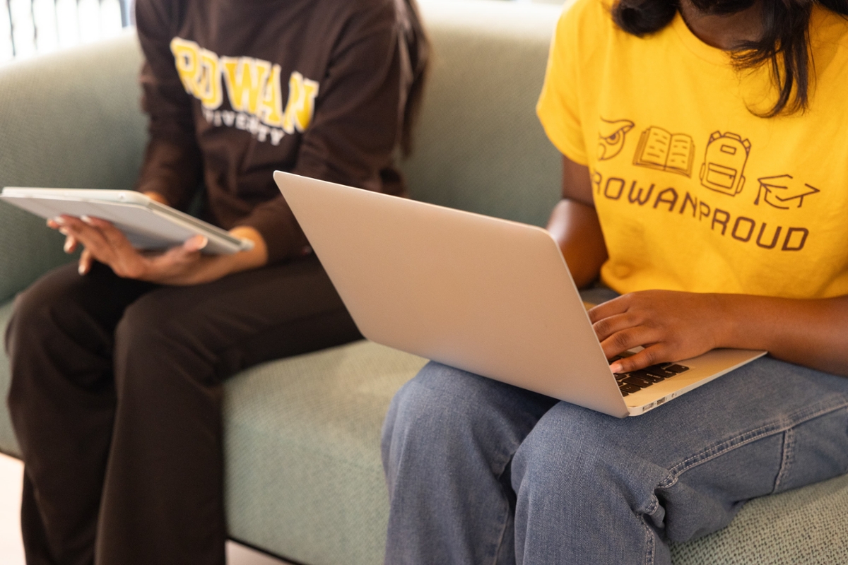 A close up image of two Rowan students next to each other. One student has a laptop on their lap while wearing a Rowan PROUD shirt and the other holding a notebook. 