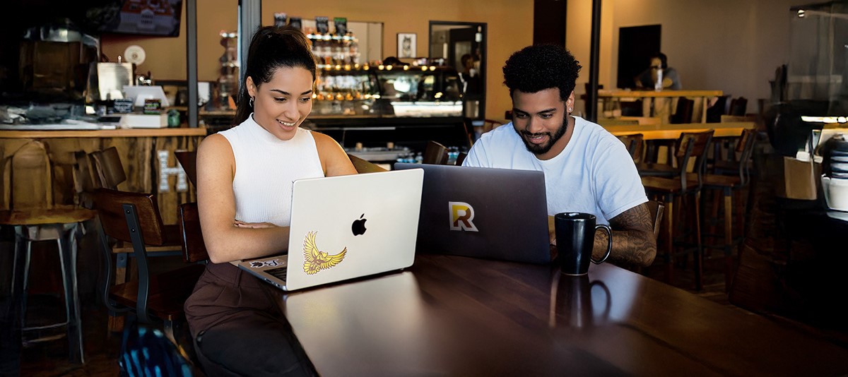 Two Rowan students sit together while working on their laptops at a cafe.