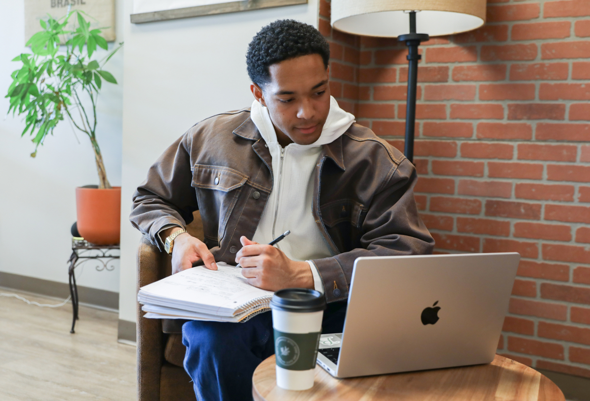 A Rowan student writing in a notebook while looking at their laptop in a cafe. 
