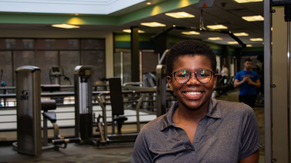 Smiling person with glasses in a gym, surrounded by exercise equipment and people working out in the background
