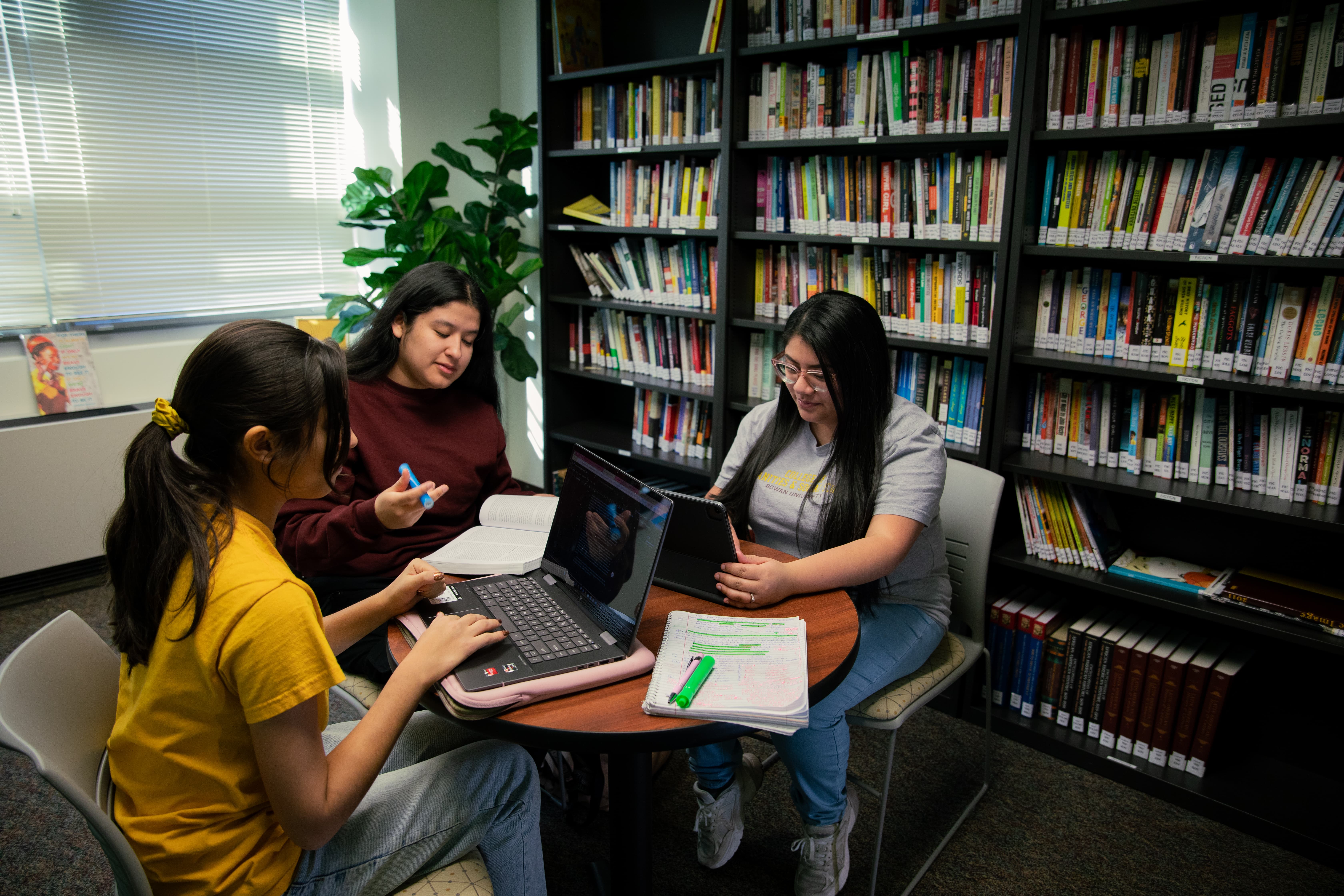 Three students sit around a table in the library while working on schoolwork.