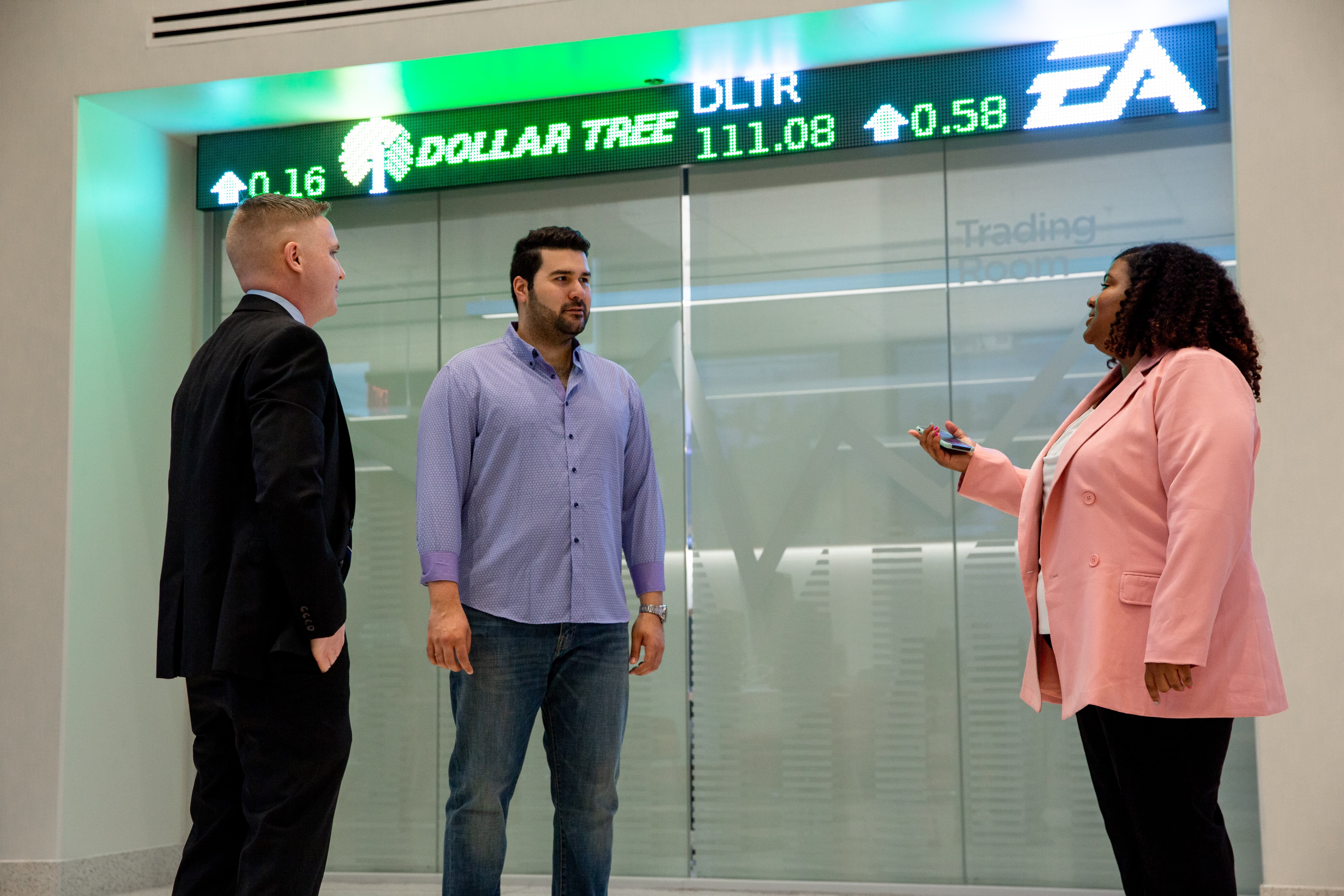 Three professionals stand while conversating in front of a stock ticker screen in the Rohrer College of Business on Rowan's main campus in Glassboro, NJ.