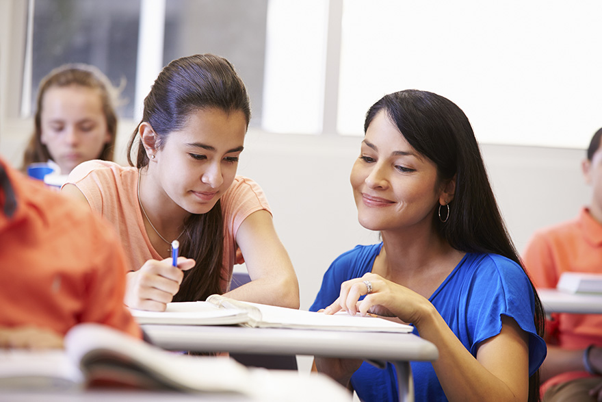 A teacher helps a student at their desk while looking at their notebook and smiling.