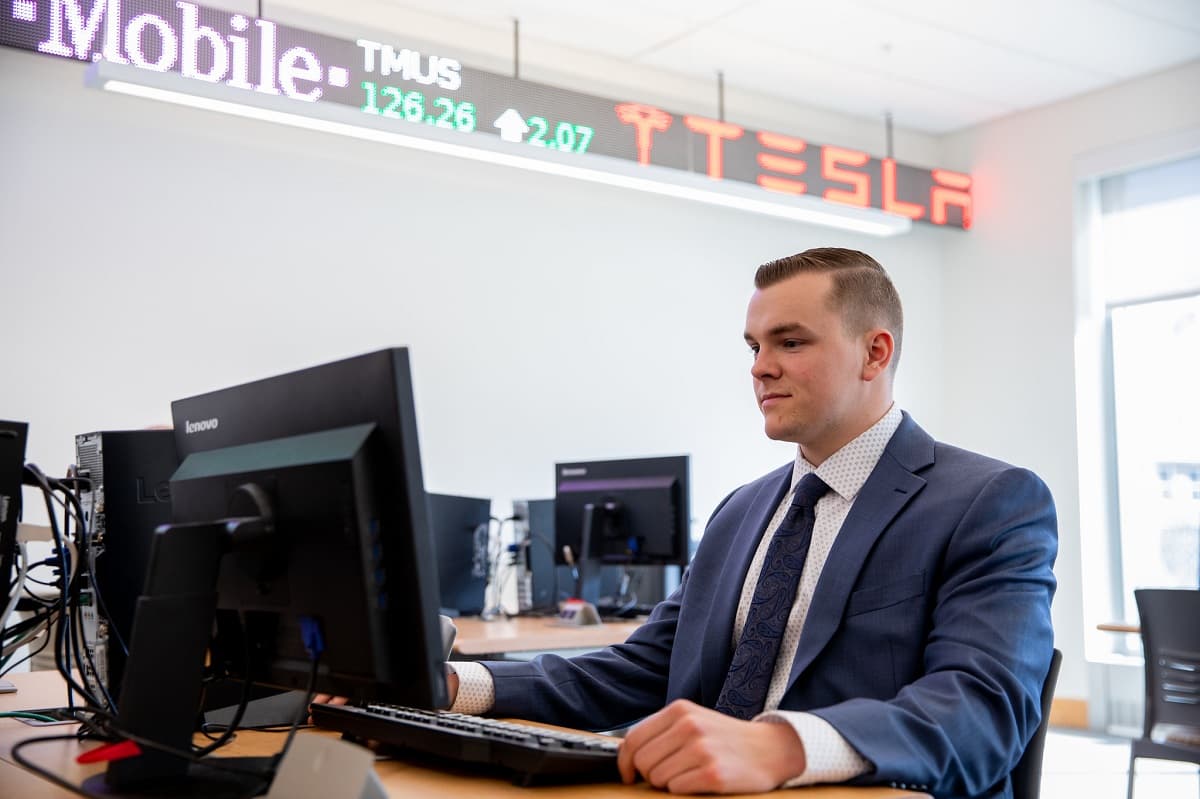 A student works at a computer with a stock market ticker overhead.