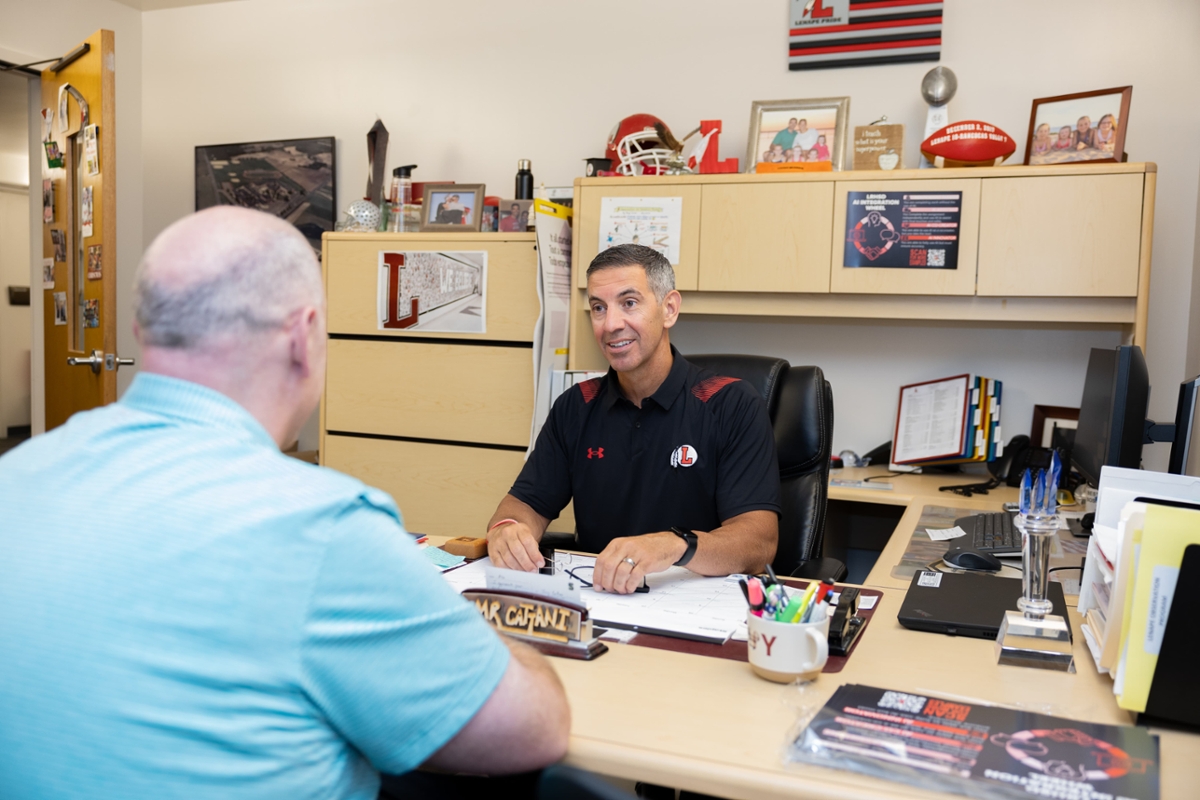 Tony Cattani, a Rowan alumni and principal at Lenape High School, sits across from a staff member while sitting at a desk.