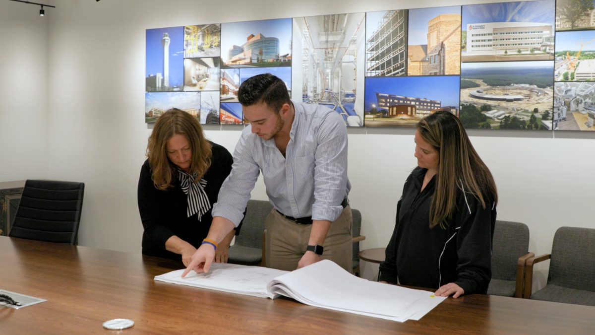 Three individuals stand in an office while looking at a construction plan on a desk in front of them.