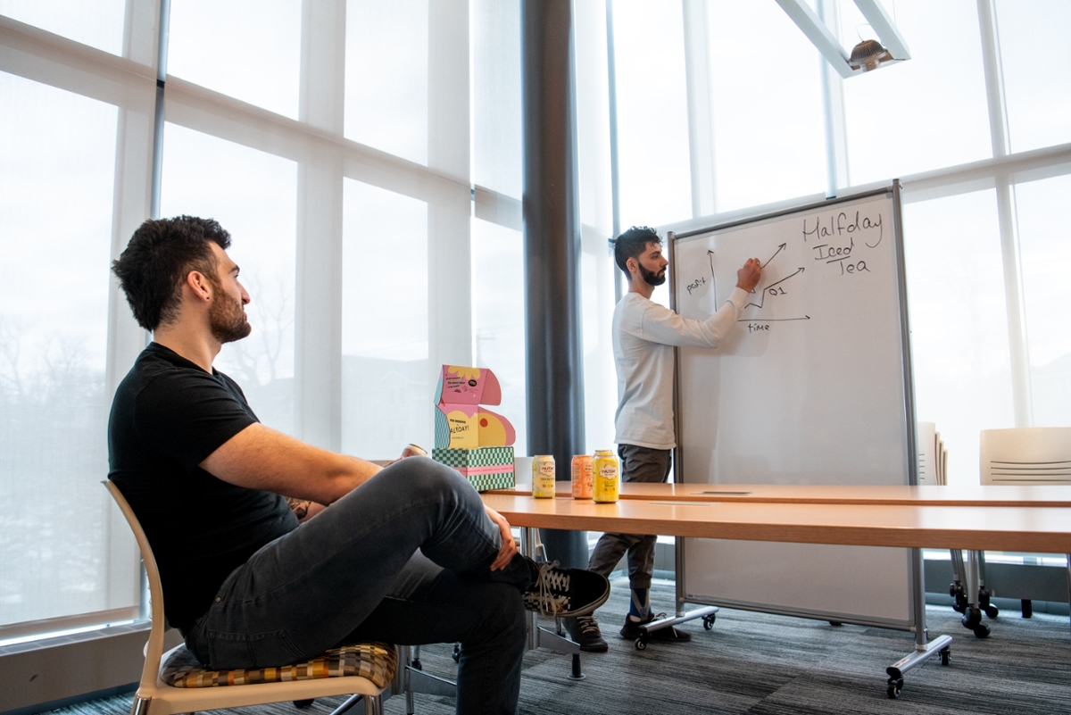 A student sits at a desk watching another student who is writing on a whiteboard in a classroom.