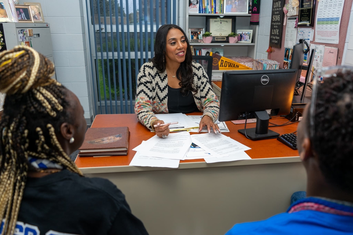 An educator sits at a desk across from students while smiling.