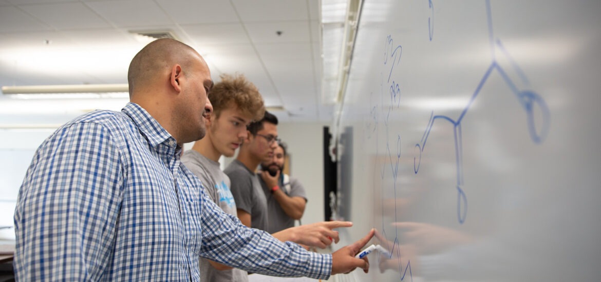 A teacher stands at the white board pointing to writing on it with students nearby.