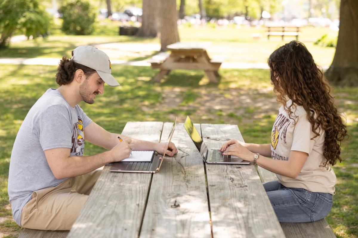 Two Rowan students sit at a picnic table while working on their laptops.