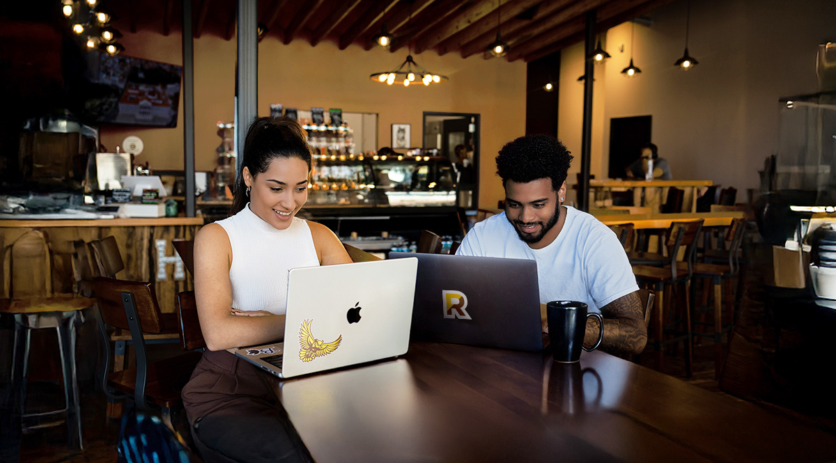 Two students smiling and looking at their laptops in a cafe in Glassboro, New Jersey.