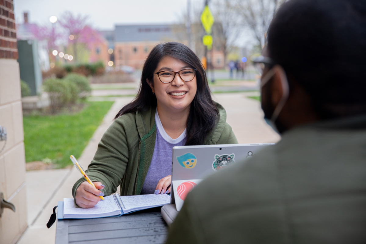 A student smiling and talking with another student while sitting with her laptop on Rowan University's main campus in Glassboro, New Jersey.