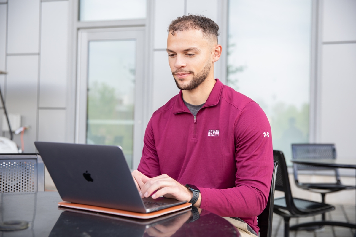 A Rowan student works on a laptop outside of a building on main campus in Glassboro, NJ.