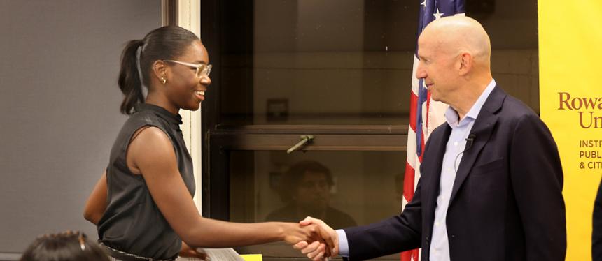 A female RIPPAC student shakes hands with former Delaware Governor Jack Markell in found of a window in the Student Center. An American flag and a yellow RIPPAC banner can be seen in the background.