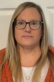 Rayna Culver smiles into the camera for a headshot while wearing an orange sweater over a white shirt, with glasses, standing in front of a white door