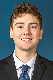 Trent Laveson smiles for a headshot while wearing a white button down shirt, blue striped tie and a grey jacket