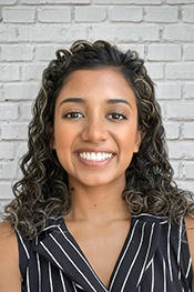 Shara Weerasuriya smiles at the camera while wearing a black and white pinstripe, sleeveless top and stands in front of a white brick wall
